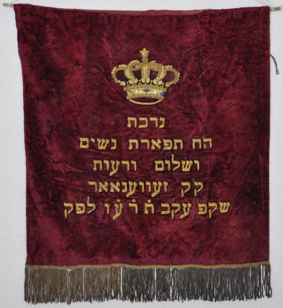 Rood fluwelen Thoramantel, met opschrift: Nedobat ha tephareth nosjim wesjaloom werowoth kk Zevenaar sj(abbat) k(oudesh) akow tod or lepok. Vertaling: Geschenk van de Vereniging Sieraad van de vrouwen en vrede en vriendschap heilige gemeente Zevenaar sabbath in het jaar 5676 (1916)